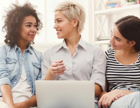 Immagine di tre ragazze a mezzo busto che chiacchierano di fronte a un computer per rappresentare come scrivere il profilo professionale del curriculum in modo efficace